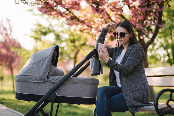 Młoda mama siedzi na ławce z dzieckiem w wózku i używa telefonu. Mothe wygląd coś w internecie - Zdjęcie, obraz