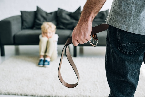 partial view of abusive father holding belt and sad son sitting on carpet - Photo, Image