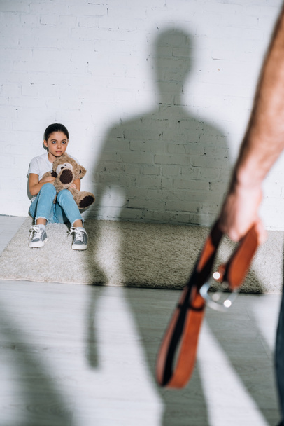 abgeschnittene Ansicht von Vater mit Gürtel und verängstigter Tochter, die auf Teppich in der Nähe seiner Silhouette sitzt - Foto, Bild