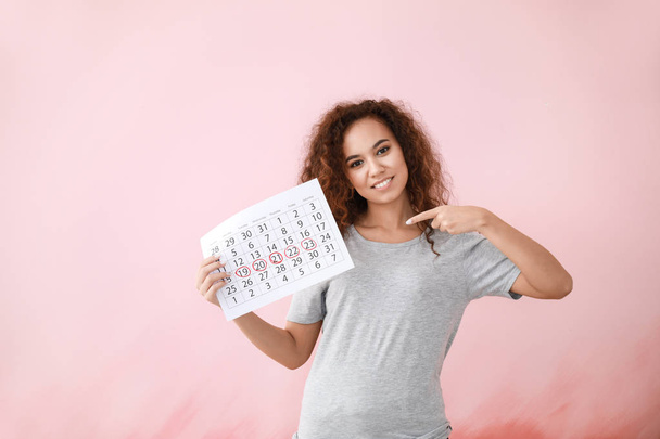 Afrikanisch-amerikanische Frau hält Kalender mit markierten Tagen der Menstruation auf farbigem Hintergrund - Foto, Bild