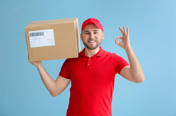 Handsome delivery man with box showing OK on color background - Photo, Image