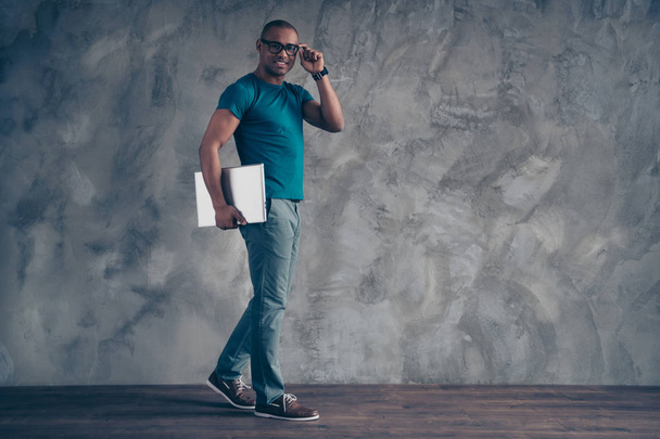 Full length body size view portrait of nice attractive good-looking cheerful cheery positive guy teacher lecturer professor in blue t-shirt carrying laptop isolated over gray industrial concrete wall - Fotó, kép