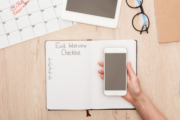 bijgesneden weergave van recruiter Holding smartphone met blanco scherm boven notebook met Exit interview checklist belettering - Foto, afbeelding