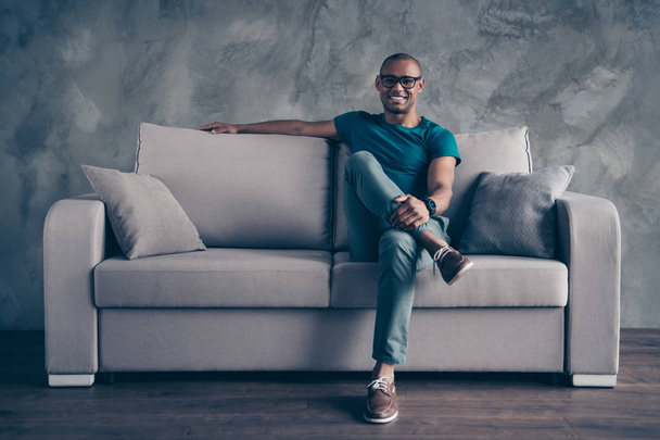 Full length body size photo amazing he him his dark skin macho hold arms hands leg pause break excited wear specs casual blue t-shirt sneakers pants sit cozy divan modern room office house indoors - 写真・画像