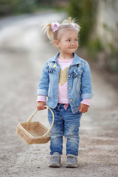 Outdoor summer portrait of a little girl in the countryside on a country road with a basket.Nature background,rural landscape,green meadow,country style.Fashionably dressed girl child with a straw basket in her hands walks alone in the park in spring - Photo, Image