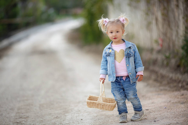 Outdoor summer portrait of a little girl in the countryside on a country road with a basket.Nature background,rural landscape,green meadow,country style.Fashionably dressed girl child with a straw basket in her hands walks alone in the park in spring - Zdjęcie, obraz