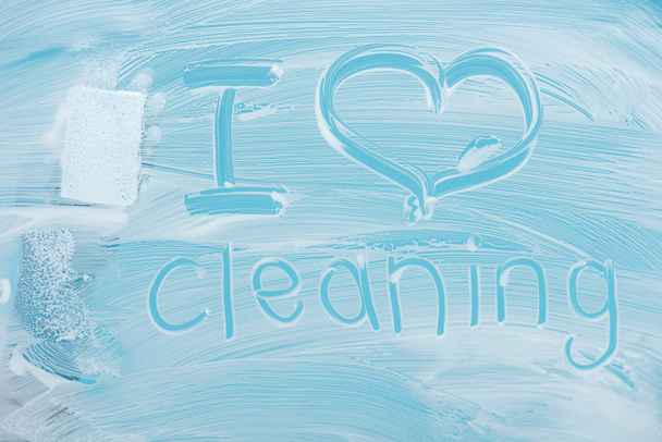 abgeschnittene Ansicht von Reiniger mit Schwamm in der Nähe Ich liebe Reinigung handgeschriebenen Schriftzug auf Glas mit weißem Schaum auf blauem Hintergrund - Foto, Bild