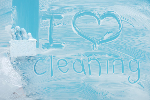 partial view of cleaner with sponge near i love cleaning handwritten lettering on glass with white foam on blue background - Foto, immagini