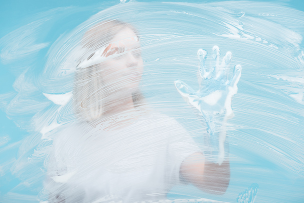 selective focus of young woman in rubber glove touching glass covered with foam on blue background - 写真・画像