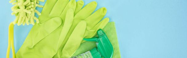 コピースペース付きの青い背景に洗剤を持つ緑色のゴム手袋、スポンジ、ぼろぼろ、スプレーボトルのパノラマショット - 写真・画像