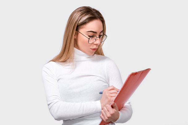 Vážná mladá žena nosí na hlavě bílou blůzu a kulaté průhledné brýle s červenou složkou v ruce a píše si nějaké poznámky. Evropská ženská pracovnice v bílém studiu. Obchodní - Fotografie, Obrázek