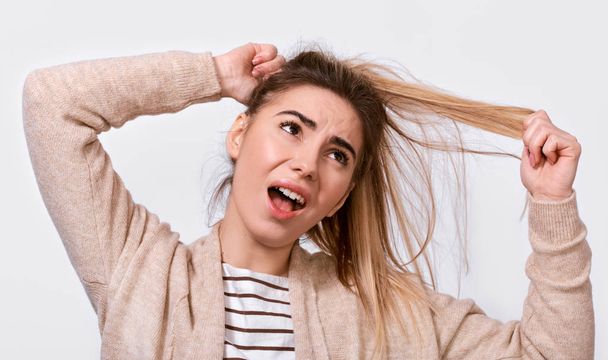 Close up image of upset young woman trying comb unruly hair pulling strands with raised hands screaming from pain and discomfort,  posing over white background. People, health and care concept - Zdjęcie, obraz
