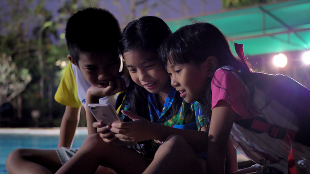 夕方にはスイミングプールの近くでリラックスしたスマートフォンを使用してアジアの子供たちのグループ. - 映像、動画