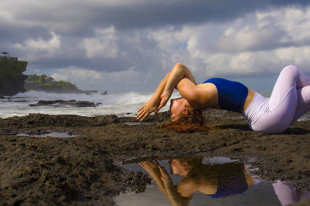 young happy attractive and beautiful red hair woman practicing yoga meditation and flexibility exercise at the beach on rocks in beautiful sea background  - Photo, image