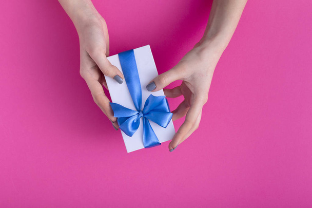 girl holding a beautiful present in hands, women with gift box with a tied blue ribbon bow in hands on a colored pink cardboard background, top view, concept holiday, love and care - Fotografie, Obrázek