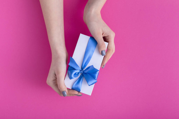 girl holding a beautiful present in hands, women with gift box with a tied blue ribbon bow in hands on a colored pink cardboard background, top view, concept holiday, love and care - Фото, зображення