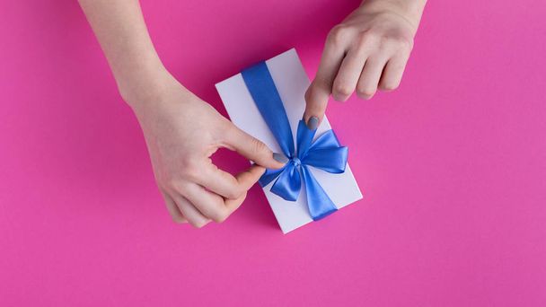 girl holding a beautiful present in hands, women with gift box with a tied blue ribbon bow in hands on a colored pink cardboard background, top view, concept holiday, love and care - Фото, изображение
