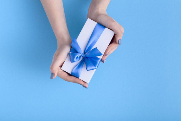girl holding a beautiful present in hands, women with gift box with a tied blue ribbon bow in hands on a pastel colored background, top view, concept holiday, love and care - Foto, immagini