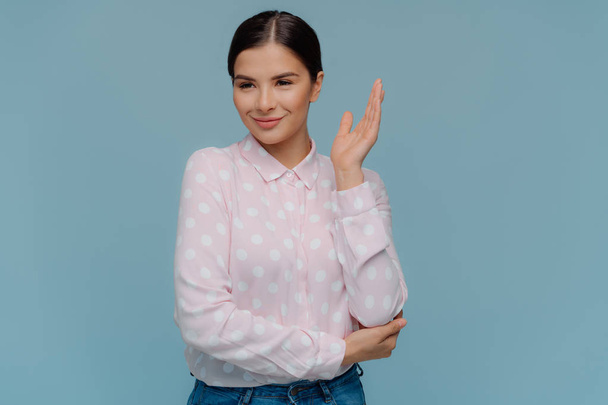 Photo of lovely young female student raises one hand, looks into distance with thoughtful expression, wears polka dot shirt and jeans, stands over blue background with blank copy space for text - Foto, Imagen