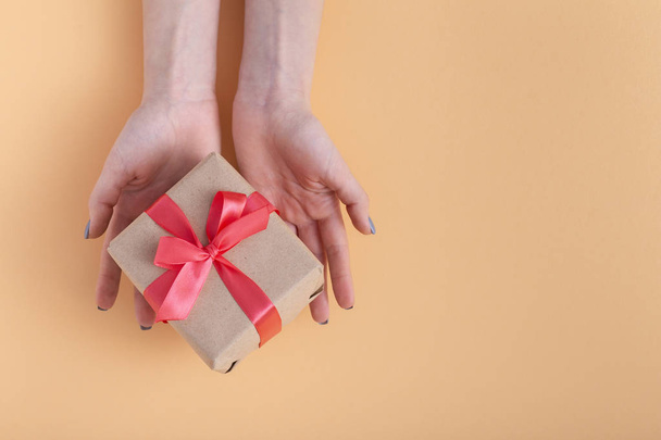 girl holding a present in hands, woman with gift box in hands wrapped in decorative craft paper with a tied red ribbon bow on a pastel colored background, top view, concept holiday, love and care - Photo, image