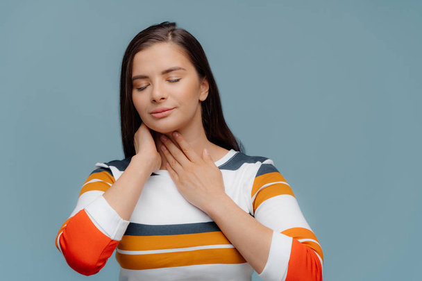 Unhealthy ill girl touches inflamated throat, suffers from pain, has eyes shut, wears casual clothing, stands indoor against blue background. Painful feelings. People and health care concept - Photo, Image