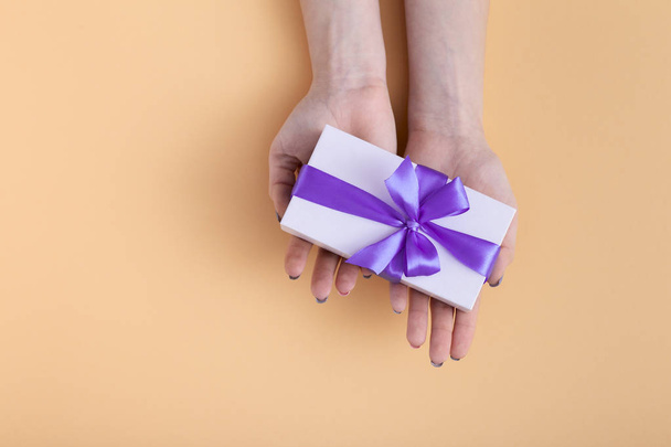 girl stretch forward on the palms present, female with gift box with a tied lilac ribbon bow in hands on a pastel colored orange background, top view, concept holiday, love and care - Fotó, kép