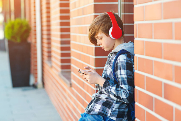 Stylish kid boy with headphones using phone at city street. Young boy plays online game at smartphone.  - Photo, Image