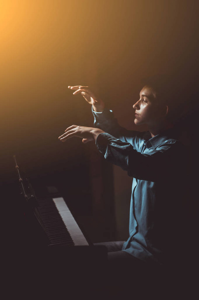 young man sitting at the piano. boy emotionally plays the keyboard instrument in the music school. student learns to play. hands pianist. black dark background. vertical - Φωτογραφία, εικόνα