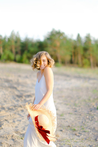 Αμερικανική ξανθιά γυναίκα περπάτημα σε βραχώδης παραλία με Hant στα χέρια και φορώντας φόρεμα. - Φωτογραφία, εικόνα