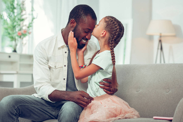 Little daughter lovingly kissing her Afro-American handsome dad forehead. - Foto, Imagem