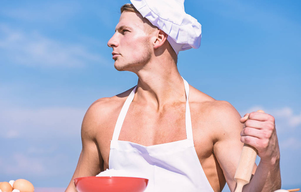 Man on confident face wears cooking hat and apron, sky on background. Cookery concept. Chef cook preparing dough for baking. Cook or chef with sexy muscular shoulders and chest holds rolling pin - Foto, imagen