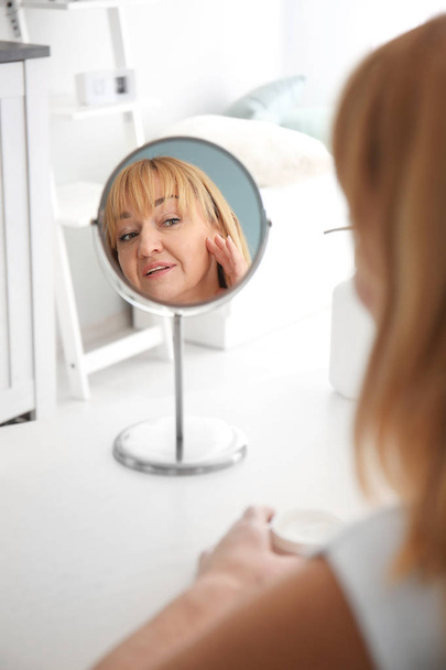 Ώριμη γυναίκα με βάζο της κρέμας κοιτάζοντας στον καθρέφτη στο σπίτι - Φωτογραφία, εικόνα