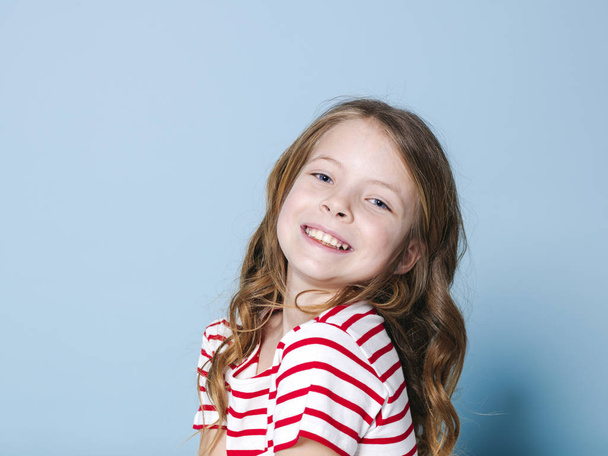 portrait of pretty girl with curly hair in striped t-shirt smiling and looking at camera while posing in front of blue background  - Zdjęcie, obraz