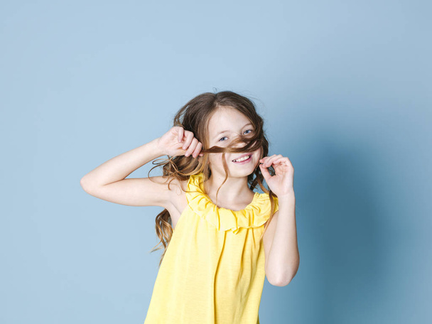 smiling pretty brunette girl in yellow top playing with hair while posing on blue background - Foto, Imagem