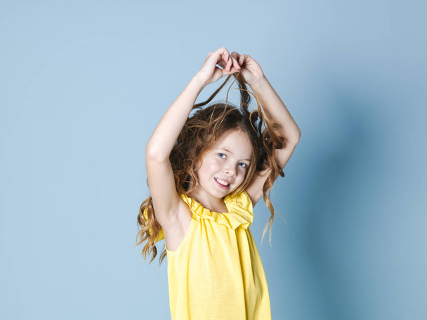 smiling pretty brunette girl in yellow top playing with hair while posing on blue background - Foto, Imagen