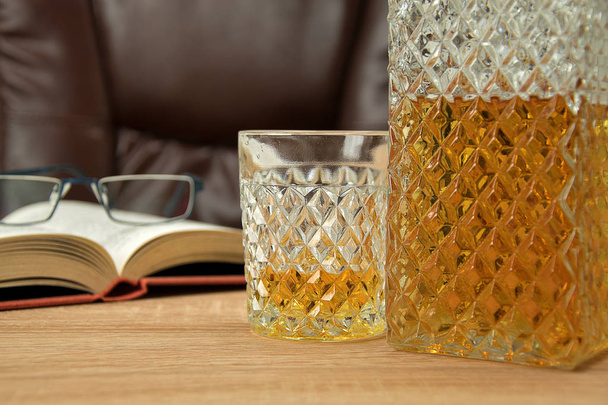 Bevanda alcolica in decanter di cristallo. In un bicchiere di whisky versato su un tavolo di legno sullo sfondo una sedia per sedersi. Libro aperto con occhiali
. - Foto, immagini