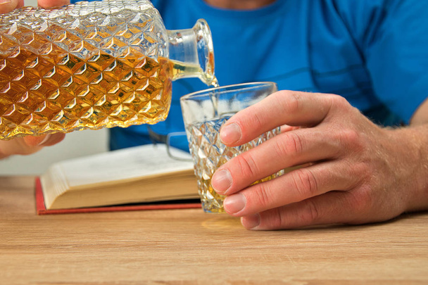 Bevanda alcolica in decanter di cristallo. Un uomo tiene un bicchiere in mano e versa un drink al whisky. Libro aperto con occhiali
. - Foto, immagini