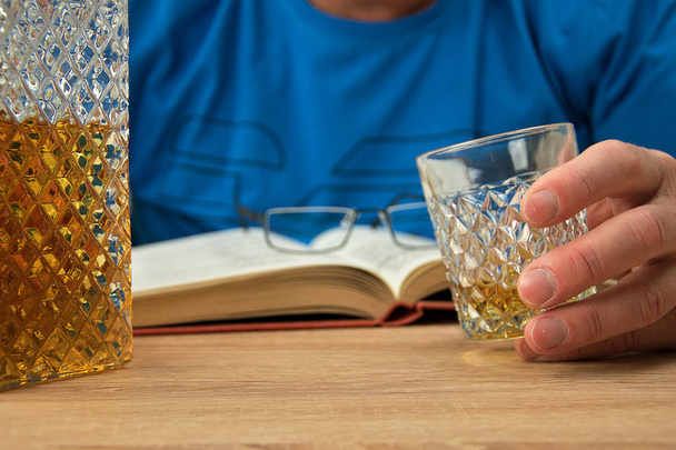 Αλκοολικό ποτό σε κρυστάλλινη καράφα. Ο άνθρωπος κρατά στο χέρι του ένα ποτήρι με ένα ποτό ουίσκι. Ανοιχτό βιβλίο με γυαλιά. - Φωτογραφία, εικόνα