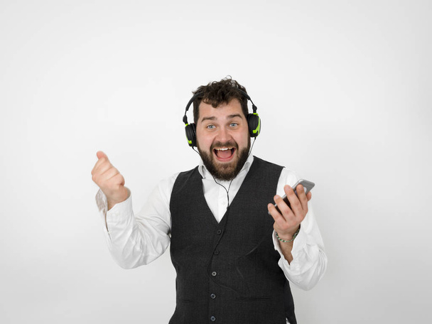 funny man with black beard listening to music in headphones and holding smartphone while posing in front of white studio wall - Photo, image