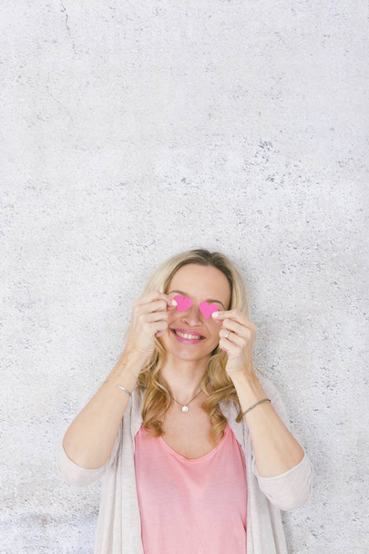funny romantic blonde woman smiling and posing with two pink paper hearts in front of grey concrete background - Photo, Image