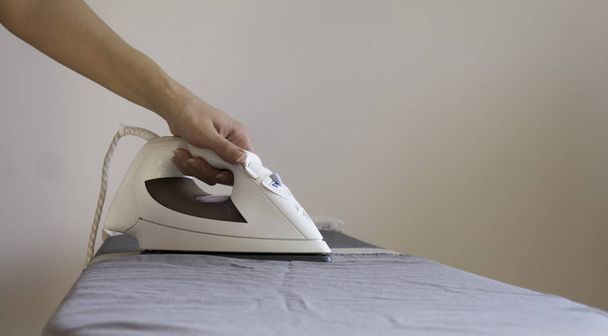 eine Hand hält ein modernes weißes elektrisches Bügeleisen bereit zum Bügeln  - Foto, Bild