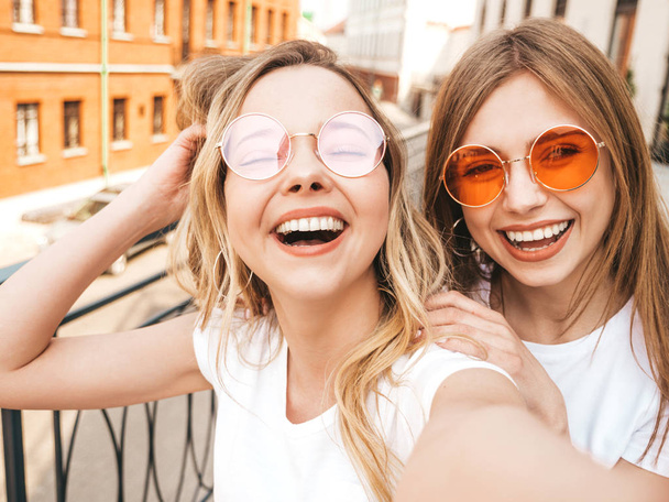 Two young smiling hipster blond women in summer white t-shirt clothes. Girls taking selfie self portrait photos on smartphone.Models posing on street background.Female showing positive face emotions - Photo, image