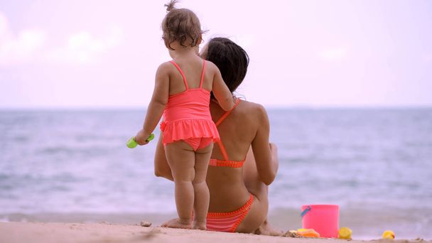 mom and daughter in pink swimsuits on the shore of a beautiful beach playing in the sand with childrens beach toys. Mom and child look into the distance on the blue sea. concept holidays. - Photo, Image