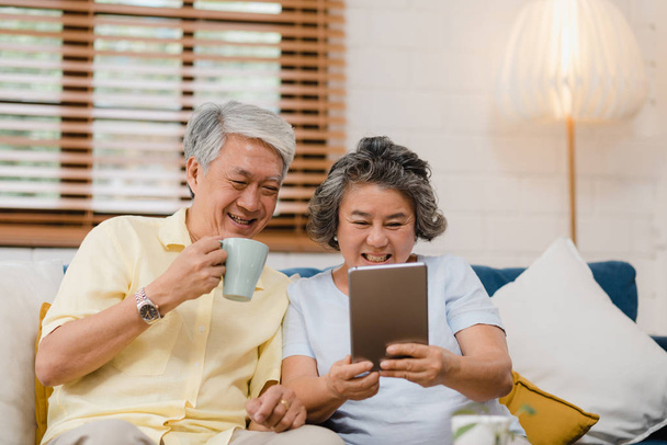 asiatisches älteres Paar, das zu Hause Tablette nimmt und Kaffee trinkt, genießt Liebesmomente, während es zu Hause entspannt auf dem Sofa liegt. Zeit genießen Lebensstil Seniorenfamilie zu Hause Konzept. - Foto, Bild