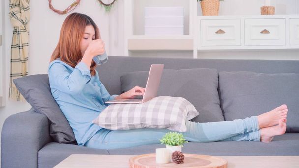 Retrato de una hermosa mujer asiática atractiva usando computadora o computadora portátil sosteniendo una cálida taza de café o té mientras está acostado en el sofá cuando se relaja en la sala de estar en casa. Estilo de vida de las mujeres en el hogar concepto
. - Foto, Imagen