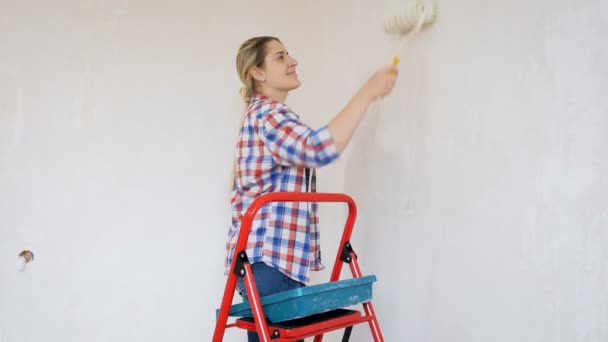Video der schönen jungen Frau im karierten Hemd bei Renovierungsarbeiten in ihrem Haus. junges Mädchen bemalt Wände mit Farbroller - Filmmaterial, Video