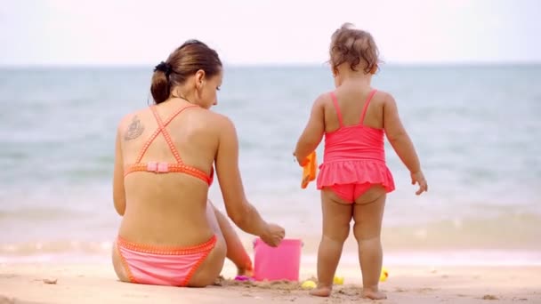 子供のビーチのおもちゃで砂の中で遊ぶ美しいビーチの海岸にピンクの水着でお母さんと娘. - 映像、動画