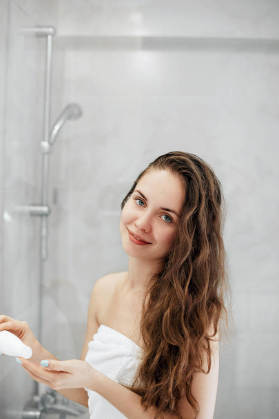 Beautiful young woman applying lotion for hair and smiling while standing in front of the mirror  in bathroom. Care hair and skin.  Girl uses protection moisturizing cream. - Fotó, kép