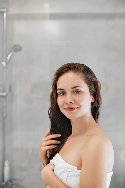 Hair and body care. Girl uses protection moisturizing cream and oil. Beautiful young woman touching her wet hair with hands and smiling while standing in front of the mirror  in bathroom.  - Zdjęcie, obraz