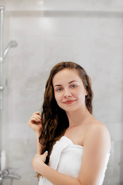 Hair. Girl touching her hair and smiling while looking in the mirror.Portrait of happy young woman with wet hair in bathroom. Care hair and skin. Girl uses protection moisturizing cream. - Фото, зображення